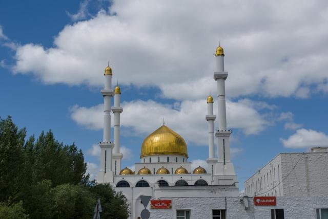 The image depicts a large, white mosque with a golden dome and four towering minarets. The mosque is situated in the center of the frame, surrounded by trees that add a natural element to the scene. In the foreground, there are buildings visible on both sides of the mosque, suggesting it may be located within an urban area or near other structures.

The sky above is clear blue with scattered clouds, providing a serene backdrop for the majestic mosque. The image does not contain any discernible text or countable objects, and the relative positions of the elements suggest that the mosque is at the center of its surroundings.