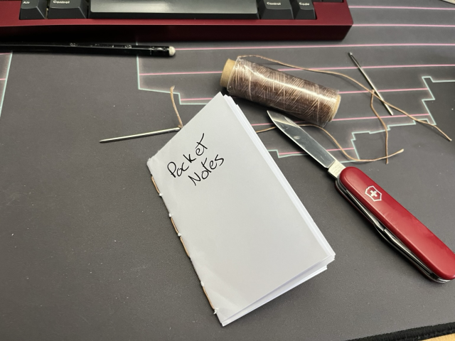 A hand sewn pocket notebook next to a sewing needle and thread and a pocketknife  
