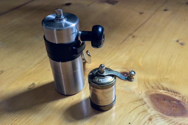 Two grinder mills on a wooden surface. One is a Porlex Mini coffe grinder. Next to it is a hand made grinder made of tin and brass at about half the size.