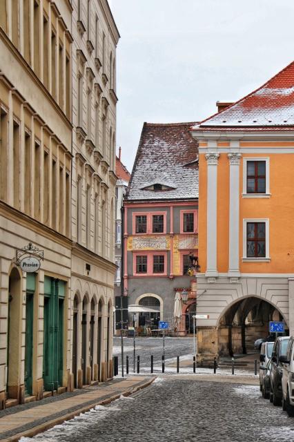 A charming cobblestone street in Görlitz, Germany, framed by historic buildings in soft pastel and earthy tones. On the left, a cream-coloured building with arched green windows displays a sign reading "Pension." At the centre, a striking pink and grey building with ornate decorations features a distinctive eyebrow dormer, giving it a whimsical, almost side-eyeing expression. To the right, a warm orange building with white accents adds a vibrant contrast. The street, lightly dusted with snow, leads toward an archway in the distance, with parked cars lining the right side.