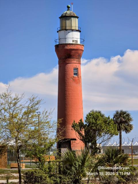 Tall red brick lighthouse with a white lantern room and a green dome top, set against a bright blue sky with scattered clouds, surrounded by greenery and a chain-link fence.