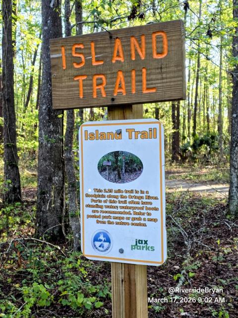 Wooden sign for Island Trail with an orange-lettered wooden sign above with details about the trail, including its length, location, and conditions, against a backdrop of trees in a park.