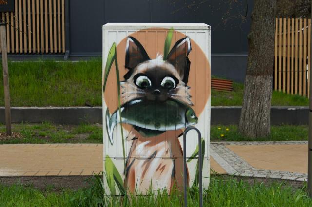 The image features a white electric box with a brown cat face and green eyes, painted on the side. The cat is holding a fish in its mouth, adding a playful element to the scene. The box is situated outdoors, surrounded by grass and trees. In the background, there are wooden fences lining up along the edge of the frame.