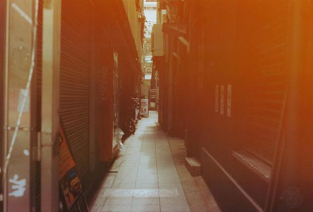 The image depicts a narrow alleyway with a warm, orange hue, due to a non-standard film processing. The alley is flanked by buildings on both sides, with closed storefronts and metal shutters. The ground is paved with light-colored tiles, and there are a few visible signs, mostly in English despite the photo was taken in Japan. The scene has a quiet, deserted atmosphere, with no people or vehicles in sight. The perspective leads the viewer's eye down the alley, creating a sense of depth.