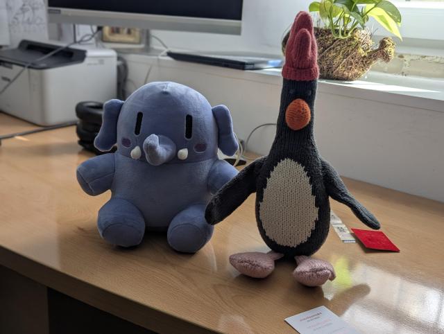 A mini Plushtodon and a Feathers McGraw plushie sat on a sunny desk in front of a window.