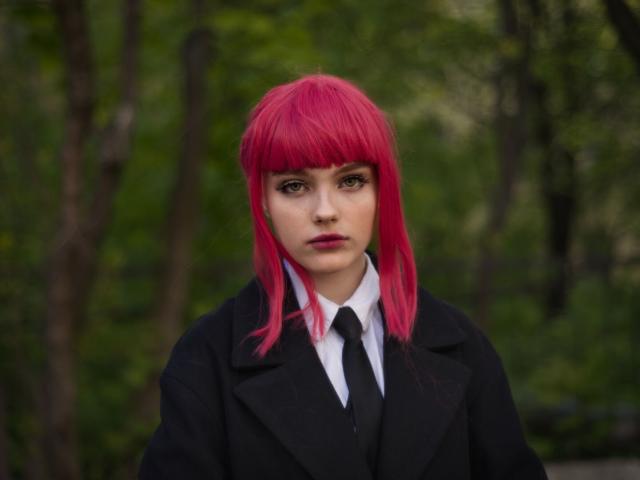 In the image, there is a young woman who is the main subject. She has vibrant pink hair that contrasts with her black attire, and green eyes. The woman is wearing a black coat over a white collared shirt with a black tie, giving her a formal appearance. She has an intense look on her face as she stares directly at the camera.

The blurred greenery in the background suggests an outdoor setting, possibly during spring or summer. The overall composition of the image is simple and focused, with the person being the central and most prominent element.