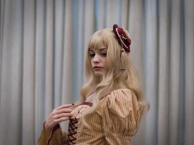 The image features a person with long, wavy blonde hair adorned with a decorative hair accessory resembling small red hat with pearls and tassels. The individual is wearing a vintage-style dress with puffed sleeves and a lace-up front, predominantly in shades of brown and cream. The person is turned slightly to the side, their expression is neutral, their right hand raised to their chest. Their posture is relaxed.

The background consists of vertical, light-colored curtains, providing a neutral backdrop that highlights the subject.
