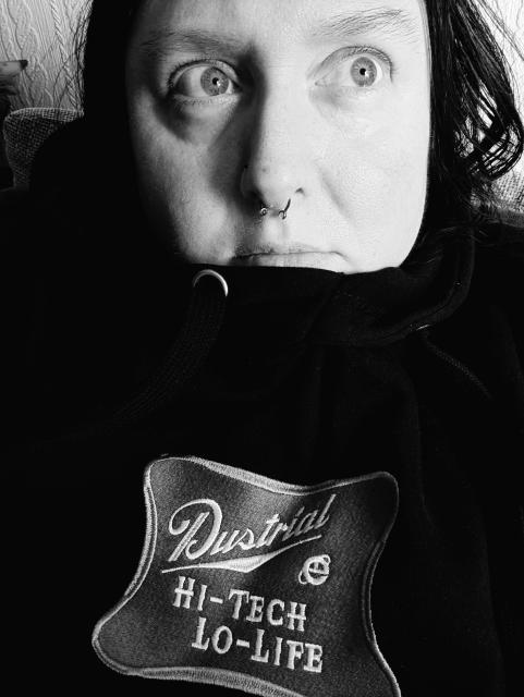 Black and white photo. A woman with dark hair and a septum piercing is looking to the side of the camera. Her chin is slightly tucked into her black hoodie which has an embroidered Dustrial patch which reads, Hi-Tech, Lo-Life.