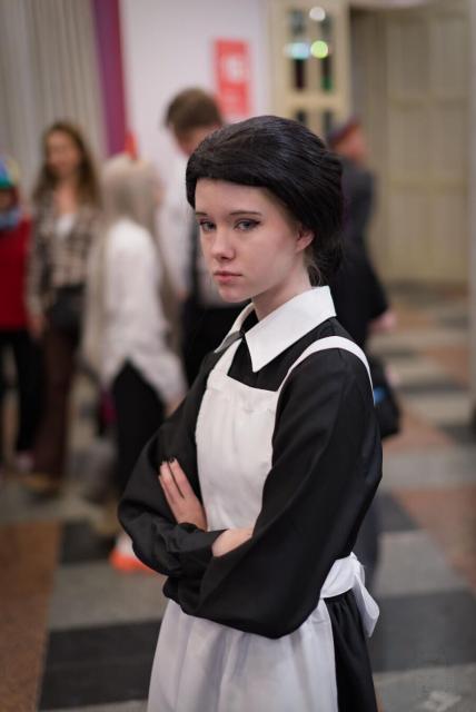 The image shows a person dressed in a maid costume, featuring a black dress with a white collar and apron. Their dark hair is styled in an updo. The individual has their arms crossed and is standing in what appears to be a public indoor space, possibly a convention or event, as there are other people in the background. The setting includes a floor with a pattern of brown, black, and gray tiles and a wall with a red sign. The focus is on the person in the maid costume, with the background slightly blurred.