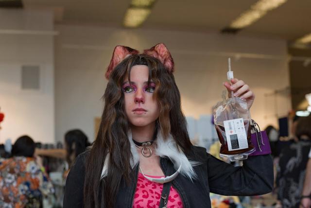 A person stands in an indoor convention hall, holding a transparent medical blood bag with a dark liquid in it, and a label containing text and a red cross. The person has long dark hair with light streaks, wolf ears (pinkish inside) attached to their head, and wears a black jacket over a pink top with a leopard print pattern. They also have a black choker with a circular pendant, and a white collar accessory around their neck. In the background, blurred figures and convention booths are visible, indicating a busy event space. The setting features overhead lighting and light-colored walls.