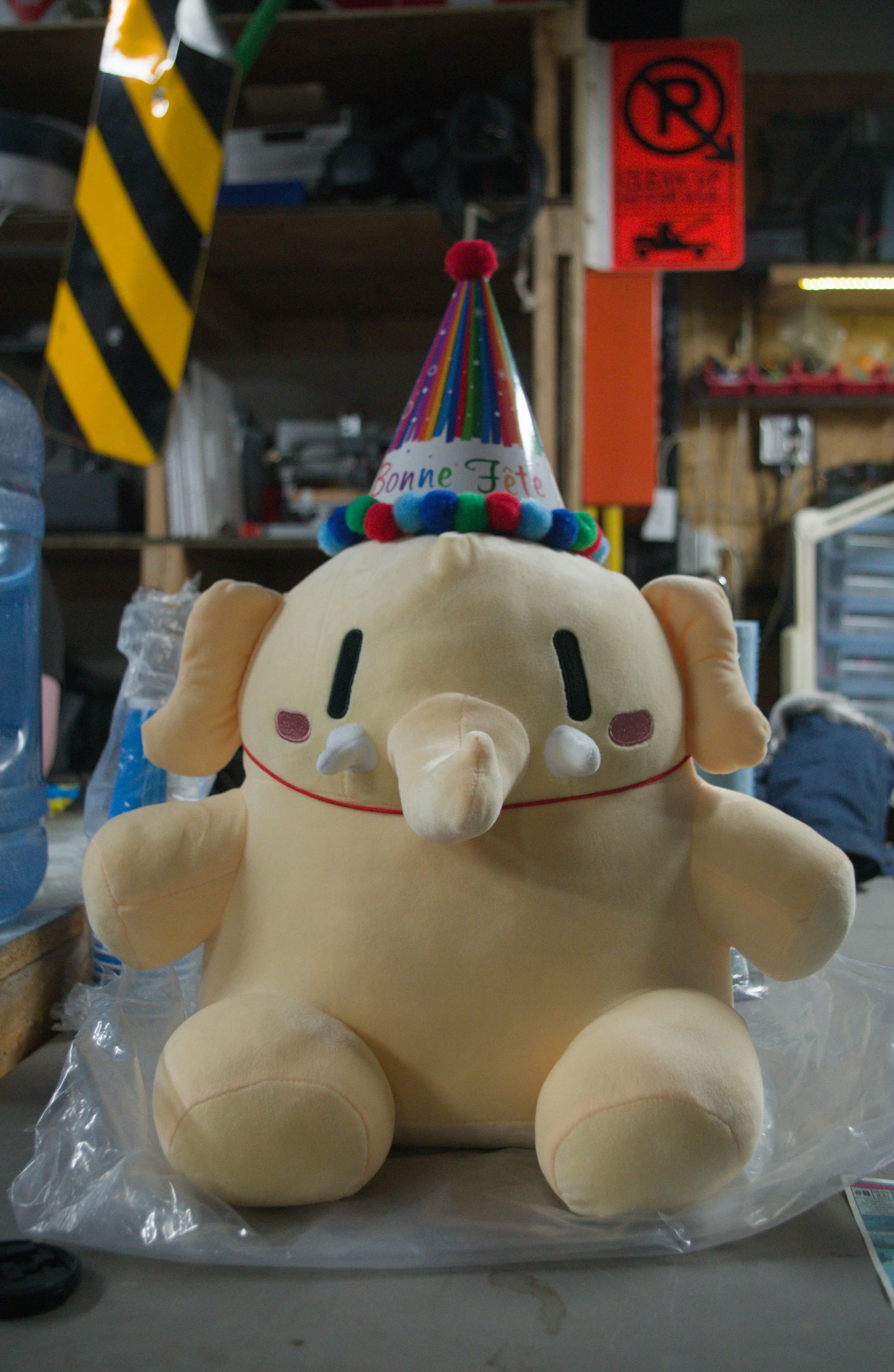 Mastodon plush wearing a birthday hat (with the French words "Bonne Fête") sitting on a table. Two traffic signs (an obstacle sign and a temporary tow-away zone sign) and shelves with various things can be seen in the background.