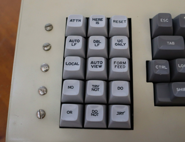 A picture of an old keyboard function pad to the left of the main keyboard. the pad contains light grey keys in a 3x5 grid. the keys are named ATTN, HERE IS, RESET, AUTO LF, DBL LF, UC ONLY, LOCAL, AUTO VIEW, FORM FEED, NO, TRY NOT, DO, OR, DO NOT and TRY with a diagonal line struck through it. To the left are flathead screws with no legends or apparent meaning. 
