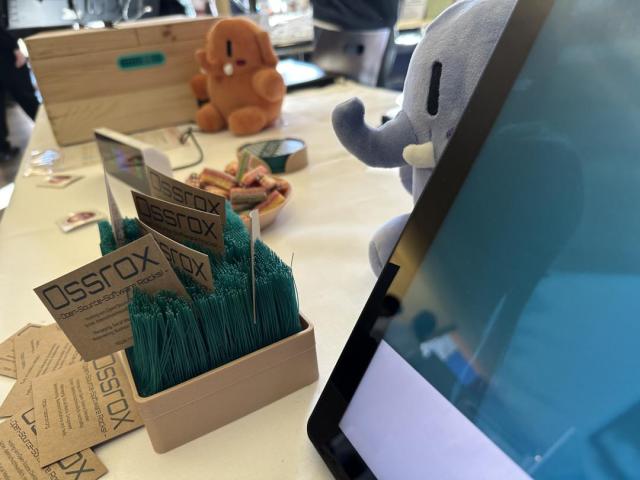 Messestand der Firma Ossrox. Auf einer weißen Tischdecke sind zwei Plushtodons (Mastodon Plüschtiere) in orange und blau zu sehen sowie diverse Visitenkarten.