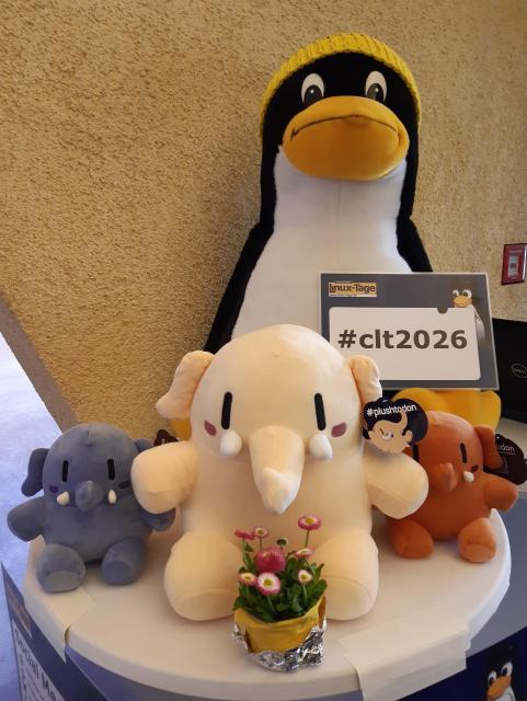 Ein großer Plüschtux und drei kleinere Plüschmastodons auf einem Tisch mit einer kleinen Blume vor sich.