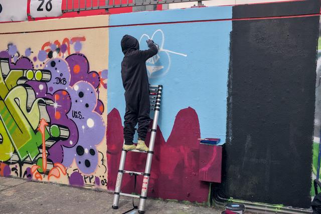 A child in a black onesie seen from behind, standing on a ladder painting with a spraycan.
