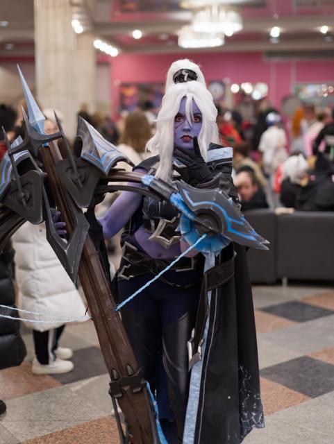 A cosplayer stands in a busy convention hall. They have long white hair, a small black headpiece, and blue face paint with red markings. Their outfit consists of a black and blue ensemble with a long black cape (trimmed in blue), dark gloves, and matching pants. They hold a very large crossbow: black with blue glowing accents, sharp angular details, and blue strings. The background shows a checkered floor (brown, black, gray tiles), pink walls, large pillars, hanging lights, and a crowd of people (some sitting on black couches). The space is well-lit with artificial lighting.