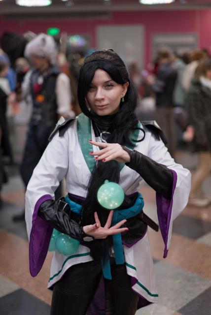 A person stands in the foreground at an indoor event. They wear a white robe with purple and teal accents, black leather sleeves, and a teal sash. The robe has green trim at the hem. They have long black hair with some strands falling over the shoulders. There is a glowing green orb apparently floating between their hands, and additional similar orbs are visible in a belt pouch. The background shows a busy room with blurred people, red walls, and overhead lighting. The scene suggests a convention or gathering.