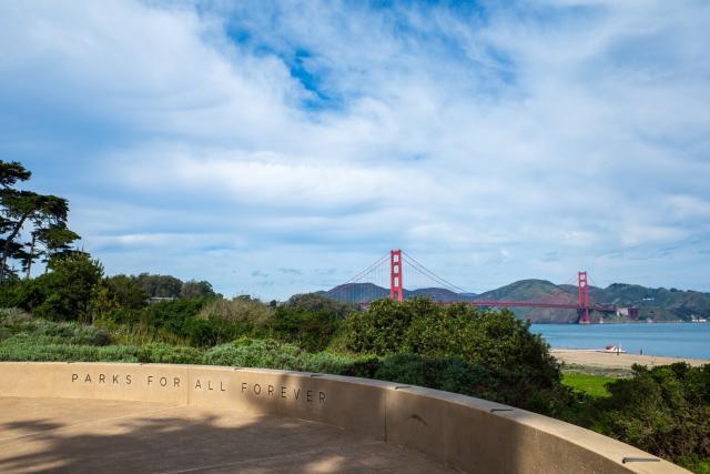 A bridge stretches across a bay behind a sign which reads "parks for all forever". [Ricoh GR III]