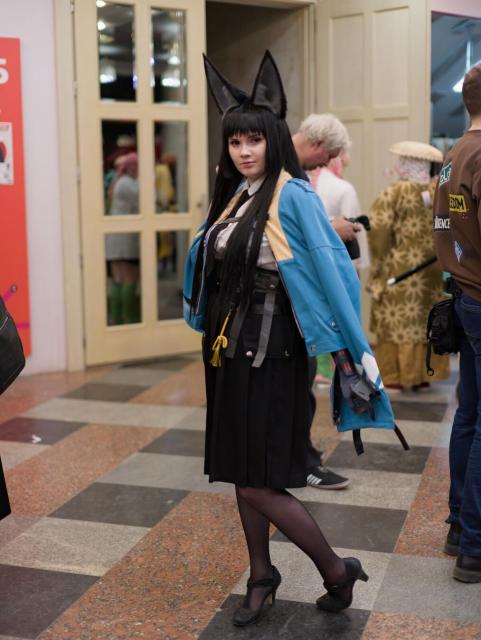 A person stands in an indoor hallway with checkered flooring (brown, gray, and beige tiles). They wear large black fox-ear headpiece, long black hair, a blue jacket with yellow shoulder accents over a white shirt and black tie, a black pleated skirt, black tights, and black heeled shoes. A black belt with gray straps and a yellow cord hangs at their waist.

In the background, there are doors with glass panels, several other people, and a wall with a part of a red panel visible. The setting appears to be a public indoor space, likely a convention or event venue.