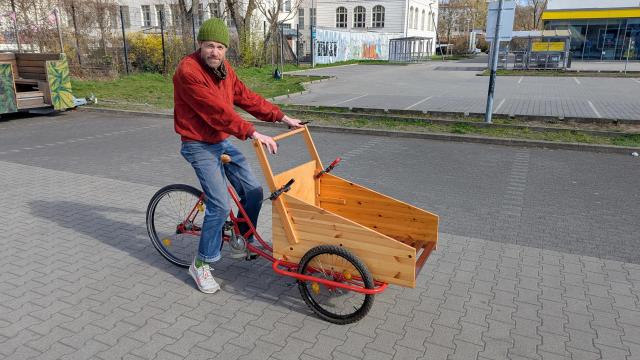 A person sitting on a 3-wheeled cargo bike in a parking lot. The handle bars are held on temporarily with clamps to a wooden cargo box.