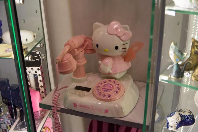 The image features a pink Hello Kitty telephone on display in a glass case. The phone is positioned at the center of the scene, with its cord extending outwards from the base to the left side of the frame. Hello Kitty herself is seated on top of the phone, her iconic bow adorning her head. Her body turned to the left towards the receiver, and her head turned to her left, towards the viewer.

Surrounding the main subject are various other objects, including some cat-like figurines to the right and something resemble to perfume bottles and packing boxes to the left, adding depth and context to the setting.