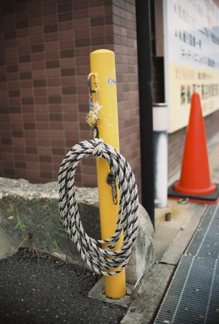 A bright yellow pole stands upright on a concrete base. A thick rope with black and white stripes is coiled around the pole, secured by metal hardware at the top. To the left of the pole is a brick wall with reddish-brown tiles. To the right, on the sidewalk, sits a red traffic cone. The sidewalk has a metal grate, and a partially visible sign with text is mounted on a dark post behind the cone. Near the pole’s base, there is dark soil or gravel and a small concrete structure.