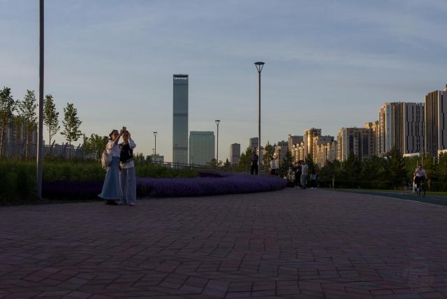 The image depicts a cityscape with tall buildings in the background, their windows reflecting the warm glow of the setting sun. The sky is painted in shades of blue and orange, creating a serene atmosphere over the urban landscape.

In the foreground, there are two individuals standing on a brick walkway, taking selfie.

Behind them, purple flowering plants add a splash of color to the scene, mirroring across a brick path that stretches into the distance. The walkway seems well-trodden, suggesting it's a popular route for park-goers or joggers.