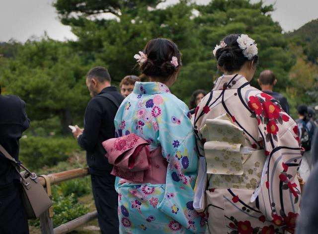 Two women in traditional Japanese kimonos stand with their backs to the camera, positioned slightly to the right of center. The woman on the left wears a light blue kimono with colorful floral patterns (pink, purple, yellow) and a pink obi (sash) around her waist. Her dark hair is styled in an updo with small pink flower accessories. The woman on the right wears a light beige kimono with red and yellow floral designs and a light yellow obi with a subtle pattern. Her dark hair is styled in an updo with white flower accessories.  

Behind them, there are people in modern clothing and a wooden railing. The background features green trees and foliage, indicating an outdoor setting like a garden or park. The scene is well-lit, suggesting daytime. The environment suggests it might be autumn due to the overcast sky and greenery that lacks vibrant summer colors.