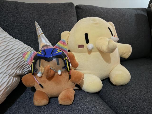 Two mastodons sitting in a sofa. One yellow and a smaller orange one. The orange mastodon is dressed with a unicorn horn and some speedy glasses. 