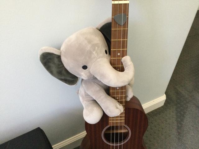 A little grey stuffed pachyderm hanging onto a ukulele.