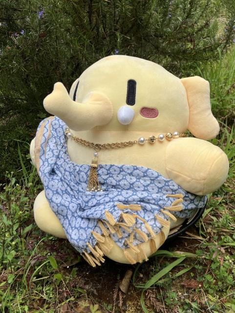 Yellow mastodon plushie, wearing a golden necklace and wrapped in a blue and gold tasseled scarf, is set under a rosemary bush.