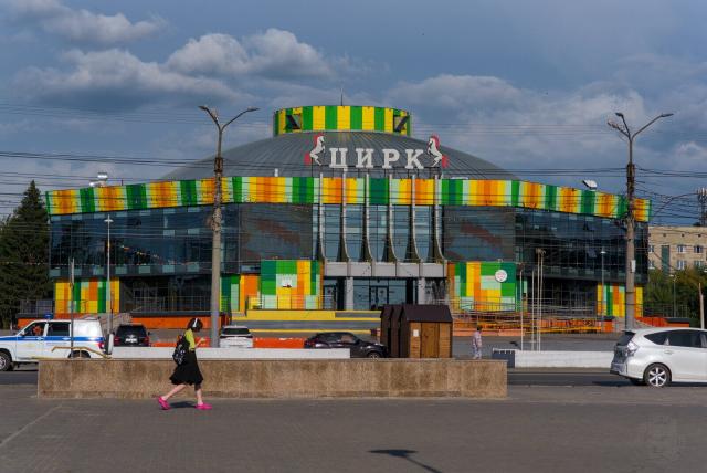 The image captures a moment on a street, where the cityscape is dominated by the large and vibrant round building. The structure stands out with its colorful stripes painted in yellow, green, and orange against a backdrop of gray skies. The large sign in Russian that translates as “Circus” is prominently displayed at the top center of the building.

In front of this architectural marvel, life goes on as usual. A woman strides confidently across the street, her pink shoes, green-yellow blouse and a backpack with lots of badges adding a pop of color to the scene. Nearby, cars are parked neatly along the side of the road, and pedestrians add life to the otherwise static urban landscape.