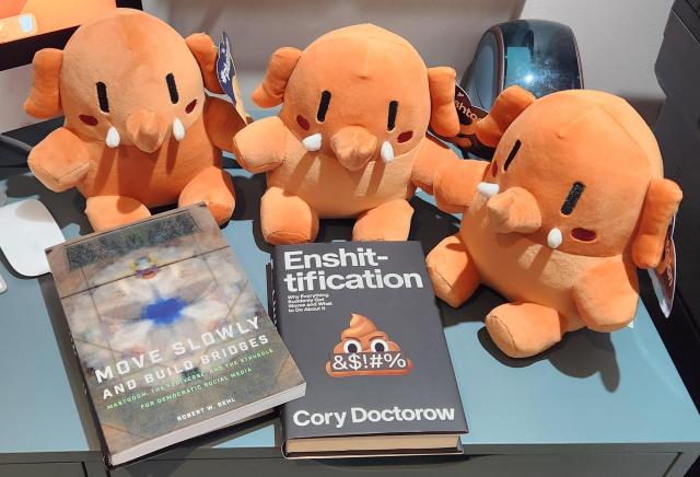 3 peach coloured Plushtodons are sitting together in a row on a desktop. In front of them are two books. The book on the left is "Move Slowly and Build Bridges" by Robert W. Gehl.  The book on the right is "Enshittification" by Cory Doctorow.