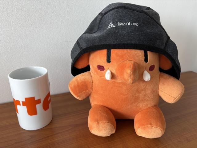 Orangenes Mastodon Plüschtier mit Mütze, die unter einen Fahrradhelm passt. Daneben in ähnlichem Farbton eine Kaffeetasse von arte