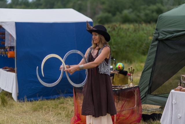 In the image, a woman is standing in front of two tents. She's wearing a brown dress and a black hat with a feather on it. The woman holds a pair of S-shaped apparently metallic forms in her hands, possibly preparing to perform an act or demonstrate some skill related to these things. Behind her, there are two blue tents set up, one larger than the other. A table is also visible near the center of the image, suggesting that this might be part of a fair or outdoor event.