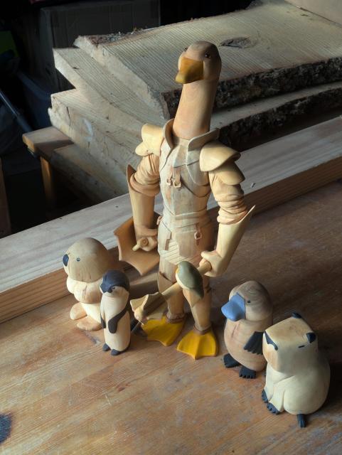 Several woodcarved animals on a workbench, a beaver, an otter, an humanoid goose wearing armor, holding a shield and a mace with robin sitting on it, a platypus and a capybara.