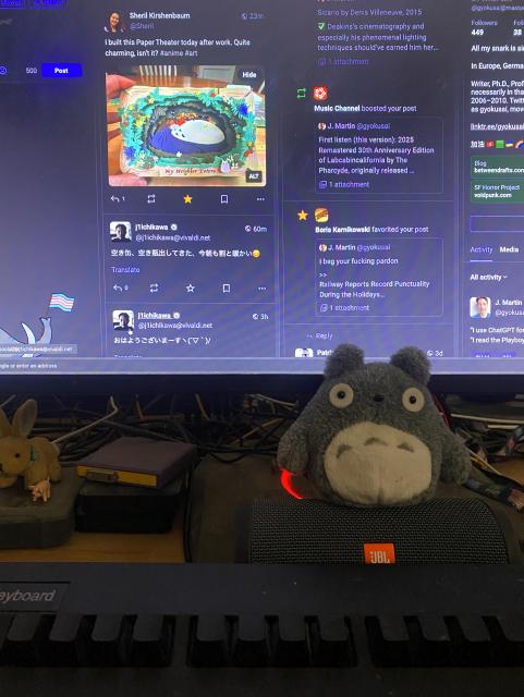 Totoro plushie sitting on a speaker in front of a screen with an open Mastodon timeline that shows Totoro artwork