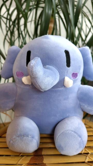 A plush mastodon, that is, a plushtodon, is looking into the camera. The plushtodon has a blueish or grayish violet body color, black rectangular eyes, pinkish violet cheeks, small white cusps, and relatively small ears. Their proboscis is slightly turned to their right. The plushtodon is sitting on a surface made of bamboo, and in the background, the leaves of a plant called "elephant's foot" (Beaucarnea recurvata) form a crown.