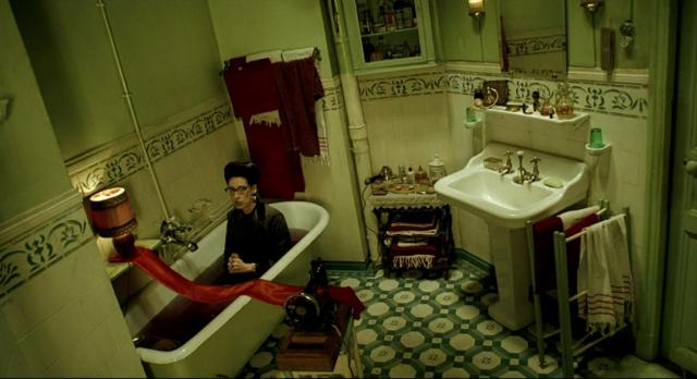 A woman, named Aurore Interligator, is sitting in a bathtub full of brown water, fully-dressed, a sewing machine is laying nearby, with a a red length of fabric running partially in the machine, then hanging over the width of the bathtub and over to a shelf, with a lit lamp sitting precariously overtop.
Aurore is looking around anxiously, hands clasped together.