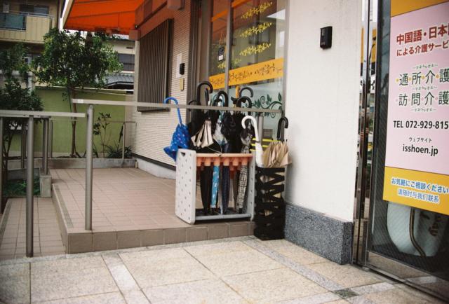 The image shows the entrance of a building with a small outdoor area. There is a rack holding several umbrellas, including blue, black, and beige ones, placed on a tiled floor. The building has a white exterior with a glass door and a signboard on the right side. The signboard contains text in Japanese and includes a telephone number and a website address. The area is partially shaded by an orange awning. There are some green plants and a small tree visible in the background. The overall setting appears to be a small business or service establishment.