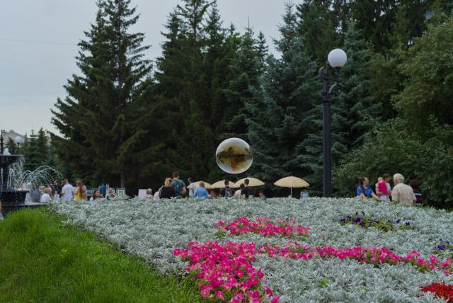 The image depicts a street scene with people walking along a boulevard and enjoying the surroundings. The visible side of boulevard is filled with trees. In the foreground there is a large flowerbed with green grass, greenish-white plants and pink and violet flowers. Above the flowerbed a large soap bubble is floating in the air, reflecting its surroundings. In the left side of the frame there is a fountain. Several umbrellas are scattered throughout the pathway in the background. People can be seen walking around, creating a lively atmosphere.