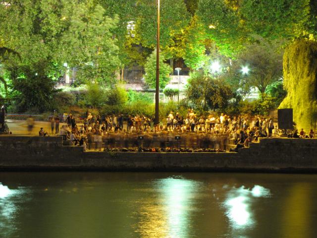 Sur les bords de seine, les gens danse, iels sont flou a cause du mouvement, tout autour d'autre gens les regardent