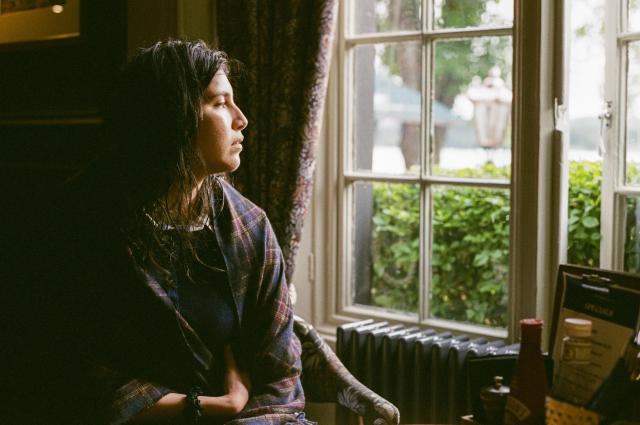 A beautiful woman with dark but wet hair sitting wrapped in a tartan shawl beside a cafe table in front of a window.