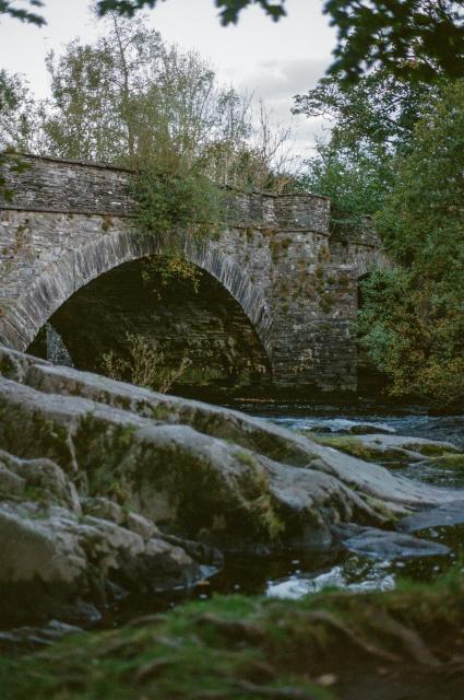 A river passing through large rocks under a stone bridge at dusk.