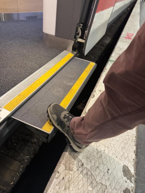 A photo of the gap filler on an DB ICE L. A matt grey platform with a yellow edge cantilevers out from the train. A right leg wearing brown trousers and a grey shoe is stepping onto the gap filler.
