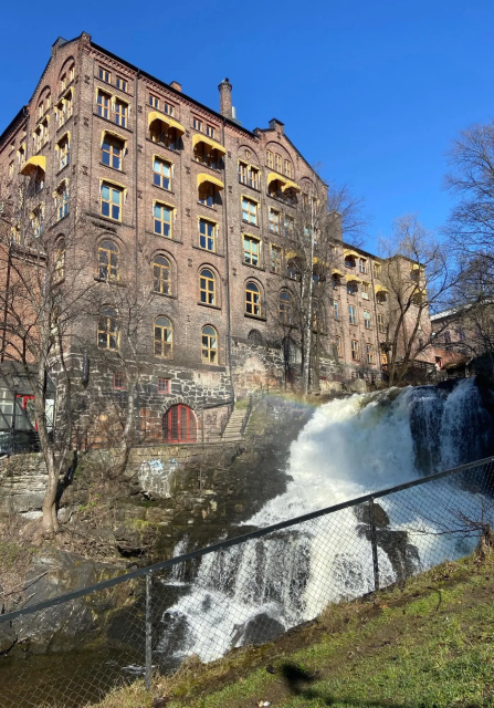Industrial brick building with yellow window frames and awnings, overseeing a waterfall. There's a rainbow on top of the waterfall.