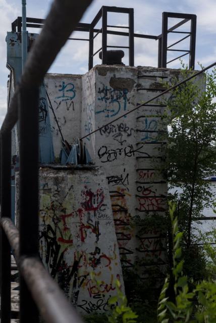 The image depicts a dilapidated concrete structure with multiple levels, a part of former river station pier. Its walls are covered in various colorful graffiti, including red, blue, green, yellow, and orange hues.

The concrete structure is situated on a river bank, with the river and some greenery on the other bank visible in the background. A metal railing can be seen near the top of the image, adding to the overall sense of abandonment.