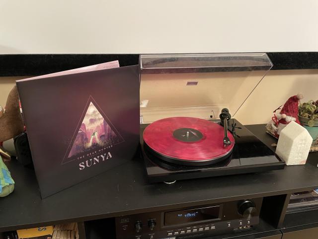 A pink translucent LP playing on a record player, next to it is the album cover with Sunya written in big letters on it. 