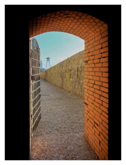 Foto im Hochformat. Blick durch einen alten Torbogen bzw.  Türbogen. Der Bogen ist aus roten Backsteinen. Alles vor dem Bogen versinkt im Schatten. Hinter dem Bogen ist eine Gittertür, die geöffnet ist, ein Weg und rechts eines Mauer. Gittertür, der obere Teil des Bogens und die Mauer Rahmen ein Stück blauen Himmel ein. Außerdem ist vor dem Himmel ein hoher Turm, der auf zwei Füßen steht und wie ein Ufo aussieht.