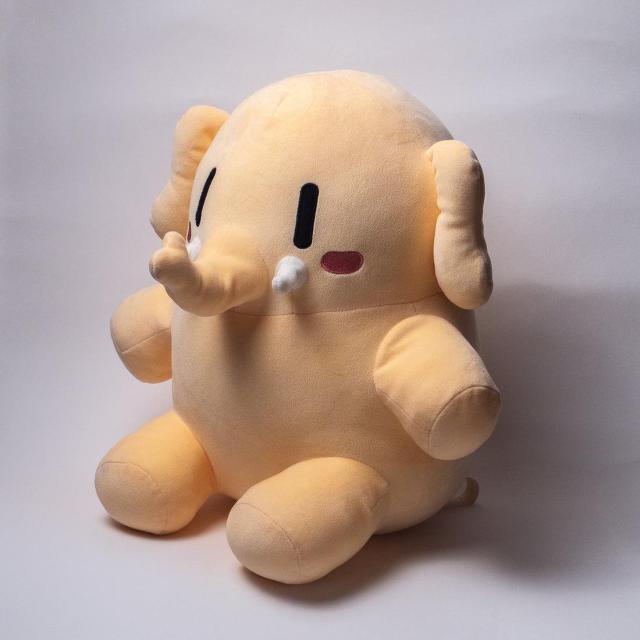 A soft, light brown elephant plush toy with a cute, cartoonish face, small black eyes, pink blush on cheeks, and a round body sits on a plain surface.